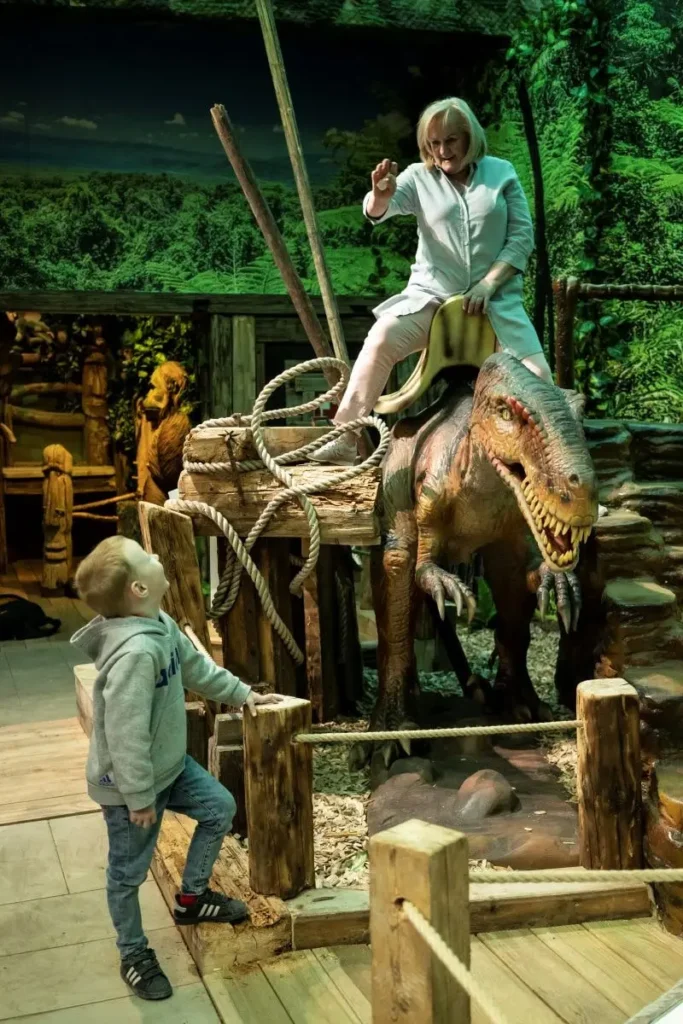 Eine Frau reitet auf einem Dinosaurier, während ein Junge staunend zusieht – eine spannende Attraktion im Tirolland beim Ausflug in Tirol.